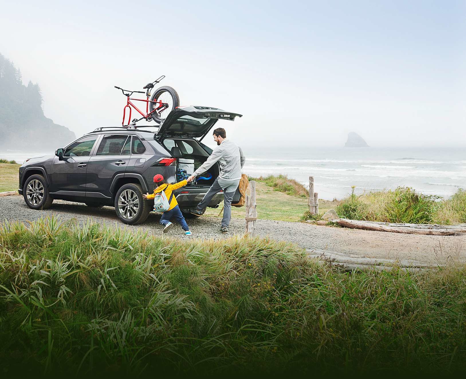 Un père et son jeune enfant debout près d'un Toyota RAV4 gris garé hors route près d'une plage par temps nuageux. Le père aide l'enfant, qui porte un sac à dos jaune. Le coffre du véhicule est ouvert et un vélo rouge est monté sur la galerie de toit.