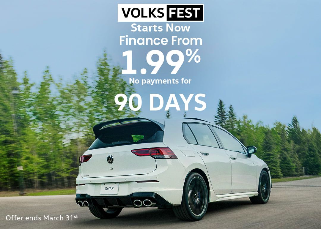 A smiling woman stands in a car dealership, looking at her smartphone while holding a takeaway coffee cup.