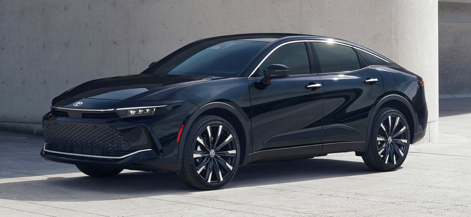 Une élégante Toyota Crown bleu foncé ou noire est garée sur une surface pavée gris clair, probablement en milieu urbain, avec un mur de béton uni en arrière-plan. La voiture est légèrement inclinée, montrant son avant et son côté.