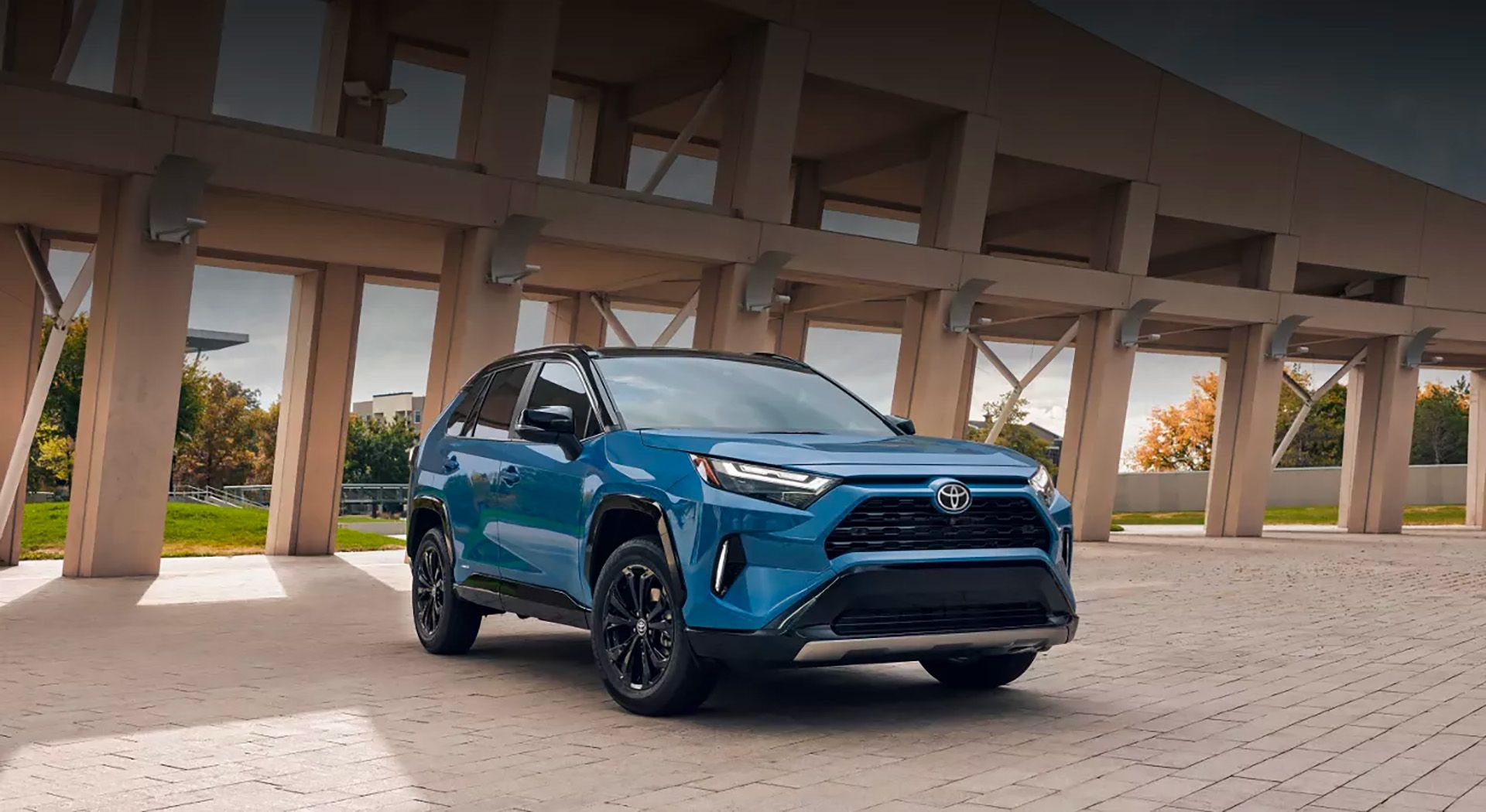 Un Toyota RAV4 bleu vif avec un toit noir et des accents noirs est garé sur une zone pavée avec des pavés de couleur claire. En arrière-plan, il y a un bâtiment moderne avec de grandes fenêtres rectangulaires et des poutres de support. Le ciel est couvert, et quelques arbres avec un feuillage d'automne sont visibles au loin.