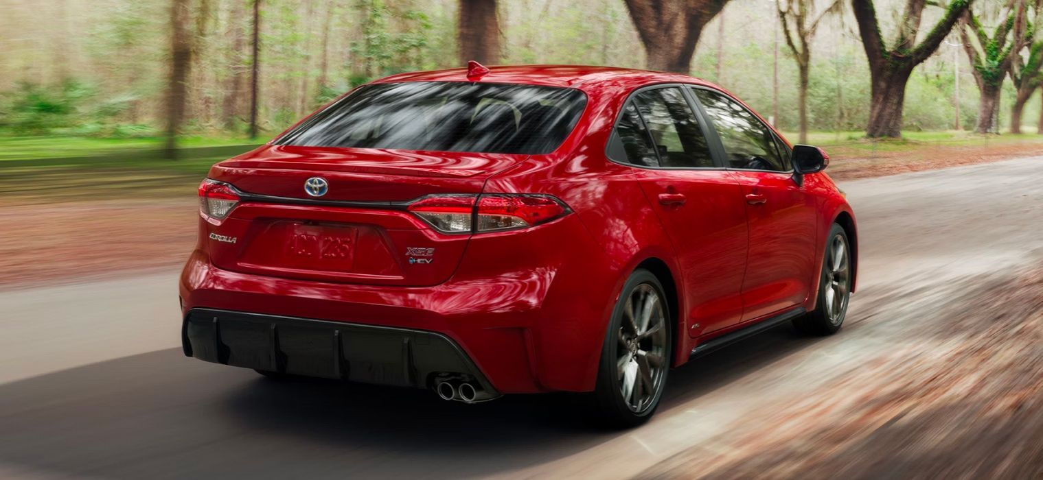 La vue arrière trois-quarts d'une Toyota Corolla Hybride rouge vif, avec l'insigne "COROLLA XSE HEV", est montrée alors qu'elle roule sur une route bordée d'arbres. Les arbres de chaque côté semblent légèrement flous, suggérant le mouvement.