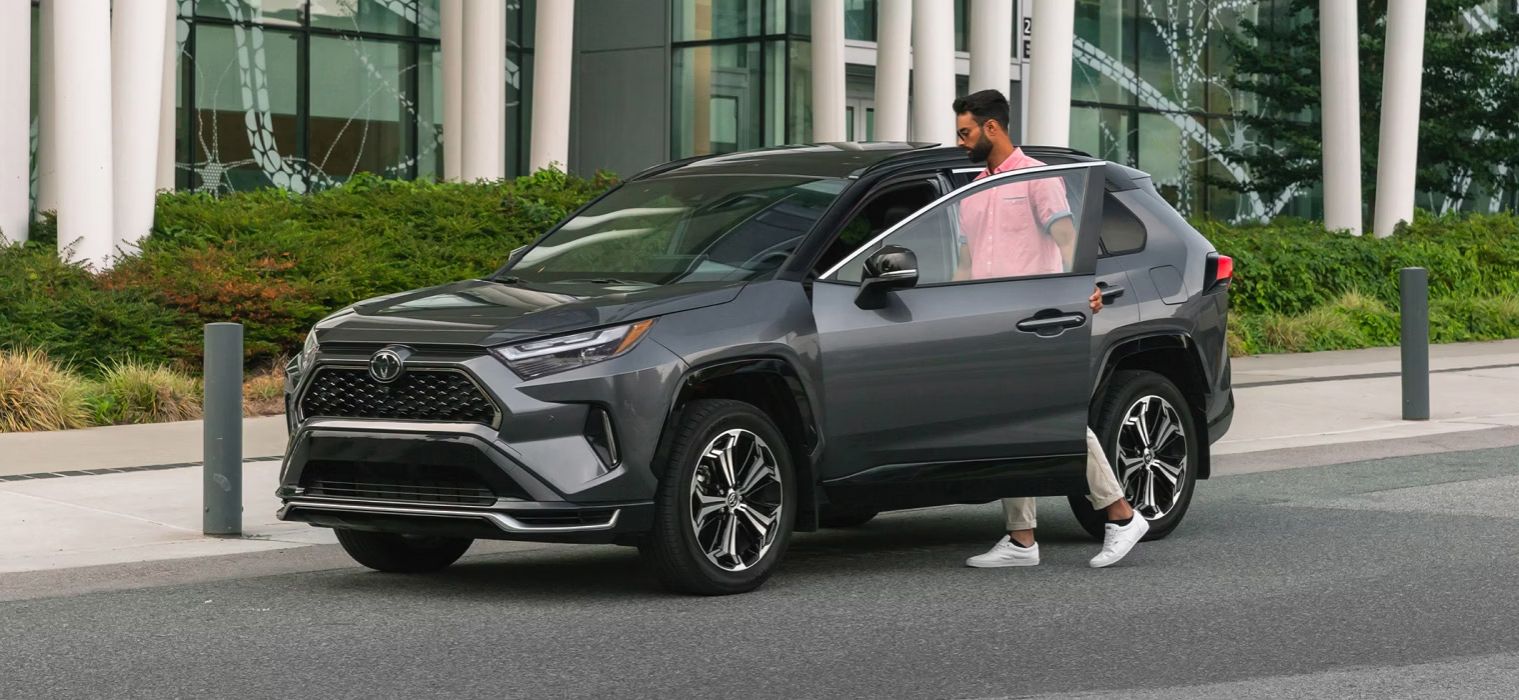 A man in a pink shirt and light pants is getting out of the driver's side of a dark gray Toyota RAV4. The car is parked on a paved surface next to a modern building with large windows and some greenery.