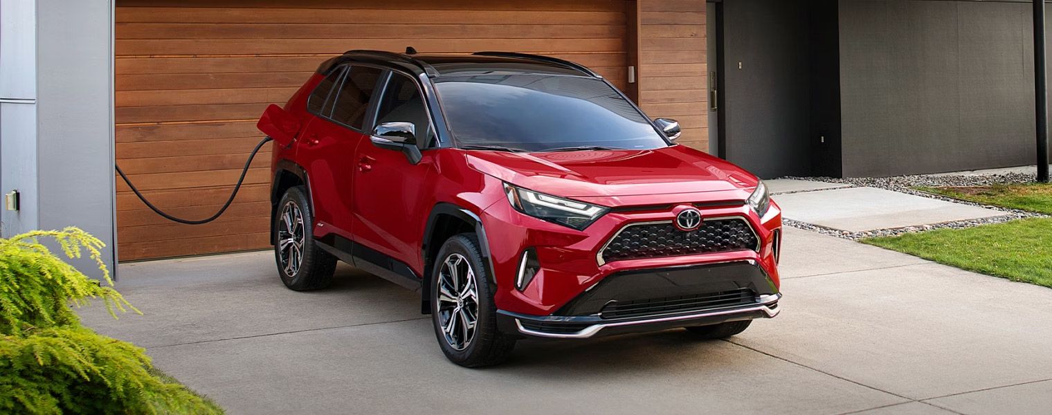A vibrant red Toyota RAV4 Prime, with a black roof, is parked in a driveway and connected to a charging station on the side of a modern house with wood paneling. Lush green foliage is visible in the foreground.