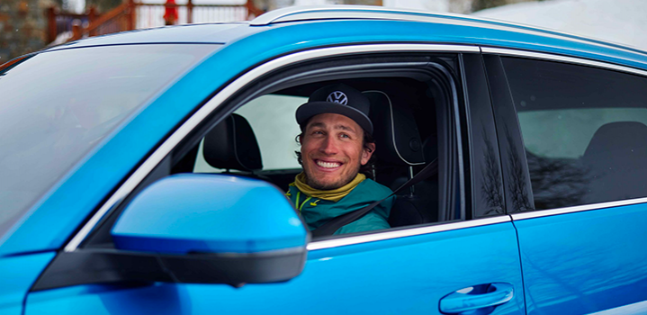 A man wearing a Volkswagen-branded hat smiles while sitting in the driver's seat of a bright blue car. He appears to be in a snowy or wintry environment.