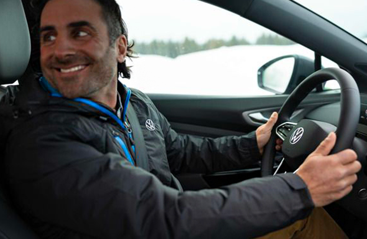 A man in a Volkswagen jacket is shown from an interior perspective, holding the steering wheel which prominently displays the VW logo. He is looking over his shoulder with a wide smile.