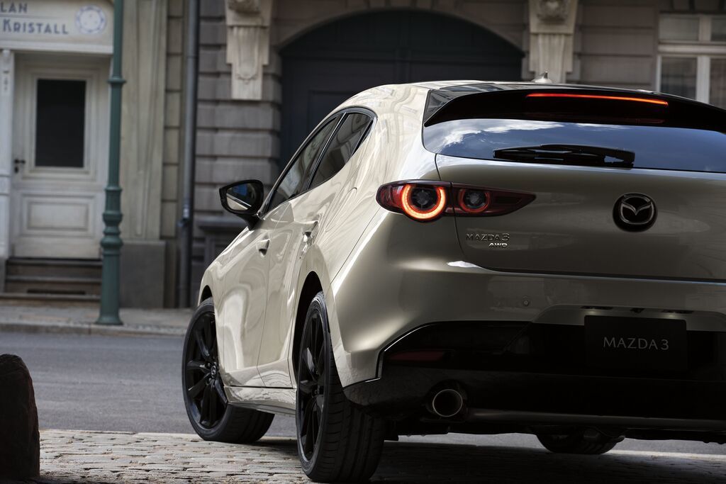 Mazda3 Sport 2025 vue arrière trois‑quarts, couleur claire, feux arrière allumés, stationnée en milieu urbain.