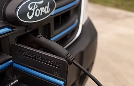 A close-up of a charging cable plugged into the front grille of a Ford E-Transit electric van.