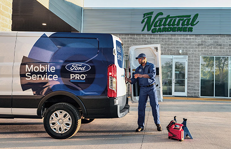 A Ford Pro Mobile Service technician working at the back of a service van parked in front of a business called "The Natural Gardener."