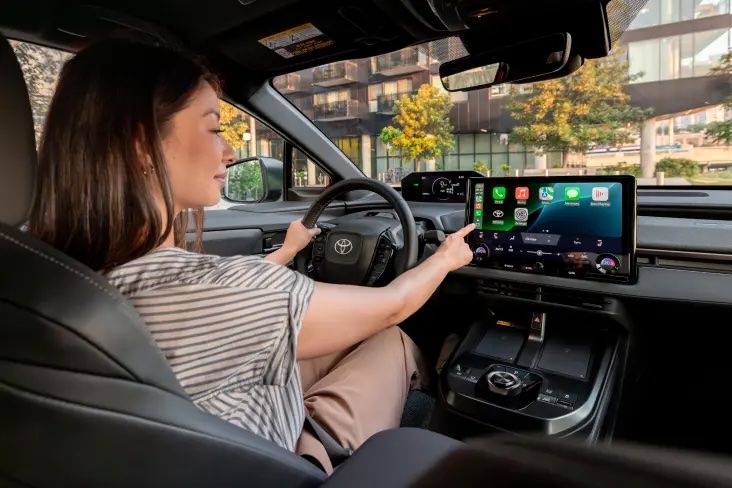 A smiling woman stands in a car dealership, looking at her smartphone while holding a takeaway coffee cup.
