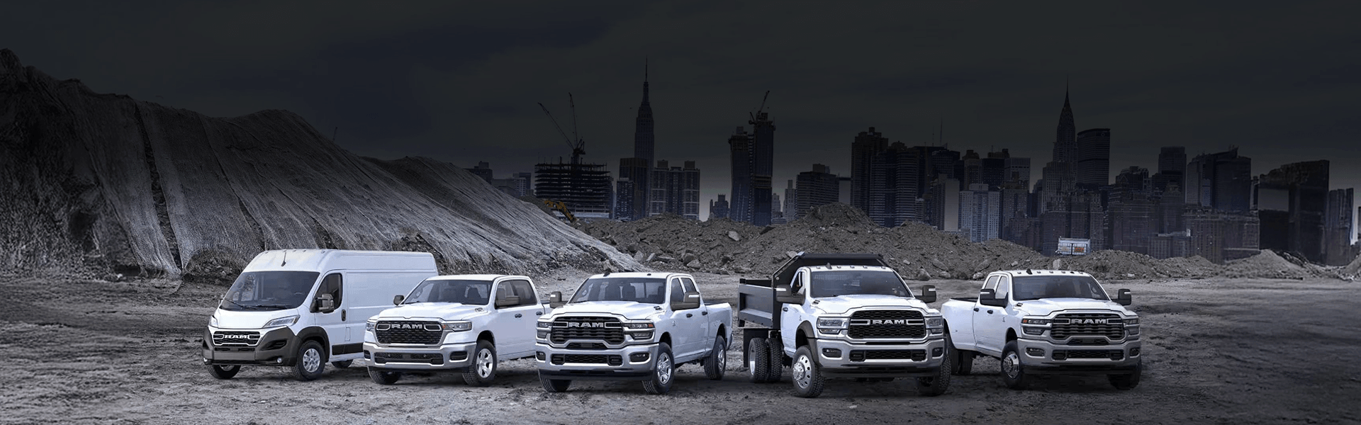 Cinq véhicules commerciaux Ram blancs, incluant une fourgonnette ProMaster et divers camions Ram, garés sur un terrain de gravier devant la silhouette d'une ville sombre.