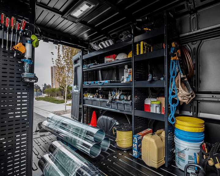 L'intérieur organisé d'une fourgonnette Ram ProMaster comprenant des étagères métalliques noires remplies d'outils, de fournitures, de bacs de rangement et de matériaux de construction.