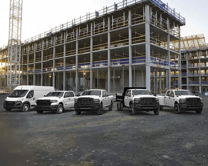 Une flotte de cinq camions commerciaux Ram blancs et une fourgonnette garés devant un grand bâtiment en béton en construction avec une grue en arrière-plan.