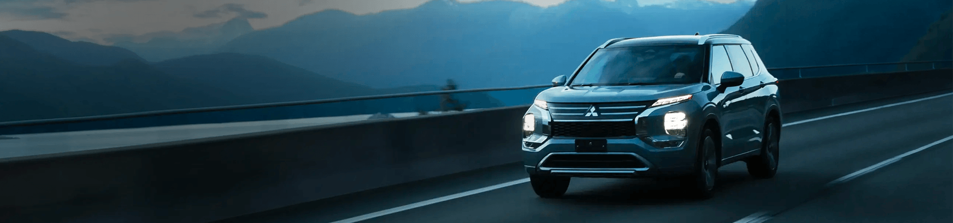 A silver Mitsubishi Outlander PHEV driving on a coastal highway at dusk, with misty blue mountains in the background.