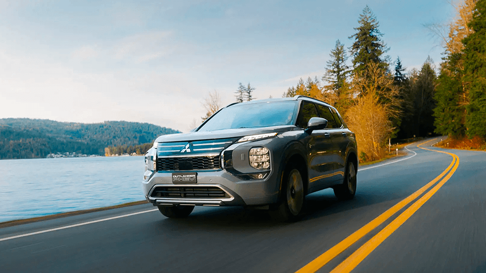 Un SUV Mitsubishi Outlander PHEV gris roulant sur une route côtière bordée de forêts.