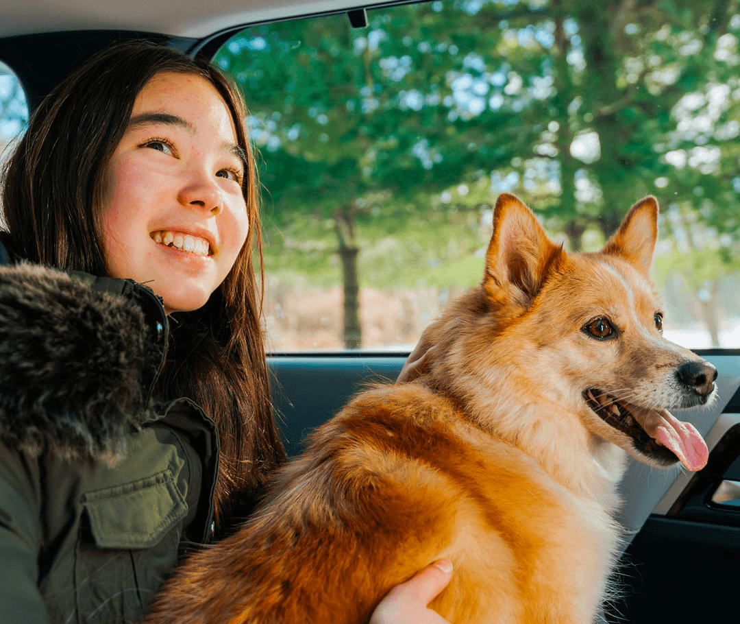 Une jeune fille souriante regardant par la fenêtre d'une voiture, assise à côté d'un chien brun clair de taille moyenne sur le siège arrière.
