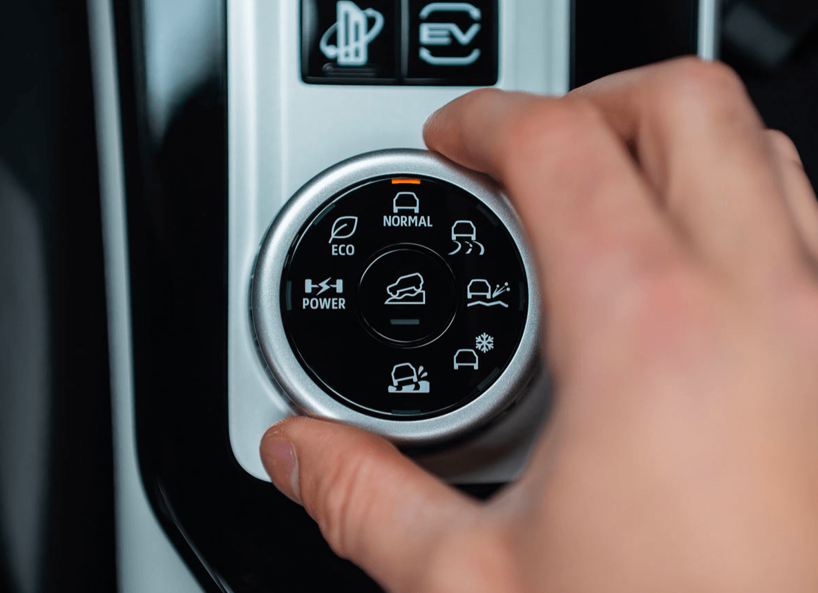 Close-up of a hand adjusting the silver rotary terrain mode selector on a vehicle’s center console, showing options like Eco, Normal, Snow, and Mud.