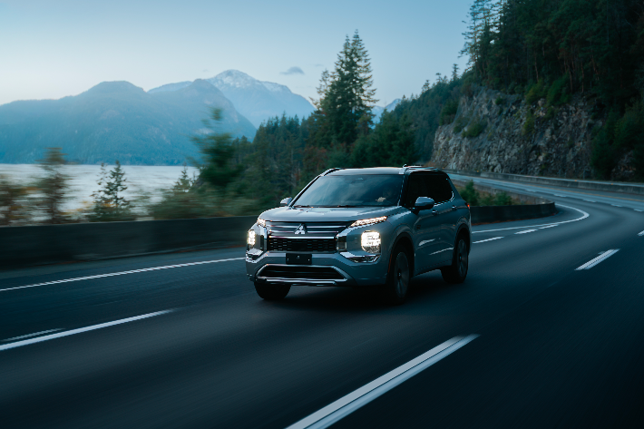 Vue avant d'un Mitsubishi Outlander PHEV argenté circulant sur une route forestière sinueuse en bordure d'un grand plan d'eau.