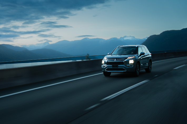 Un Mitsubishi Outlander PHEV argenté circulant sur une autoroute côtière au crépuscule, avec des montagnes bleues brumeuses en arrière-plan.