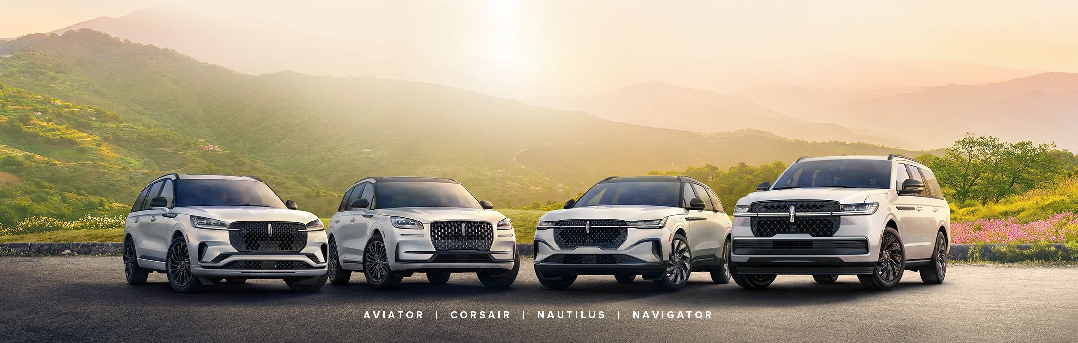 Four white Lincoln SUVs, from left to right, the Aviator, Corsair, Nautilus, and Navigator, are parked side-by-side on a road. In the background, lush green mountains rise under a bright, sunny sky with warm light.
