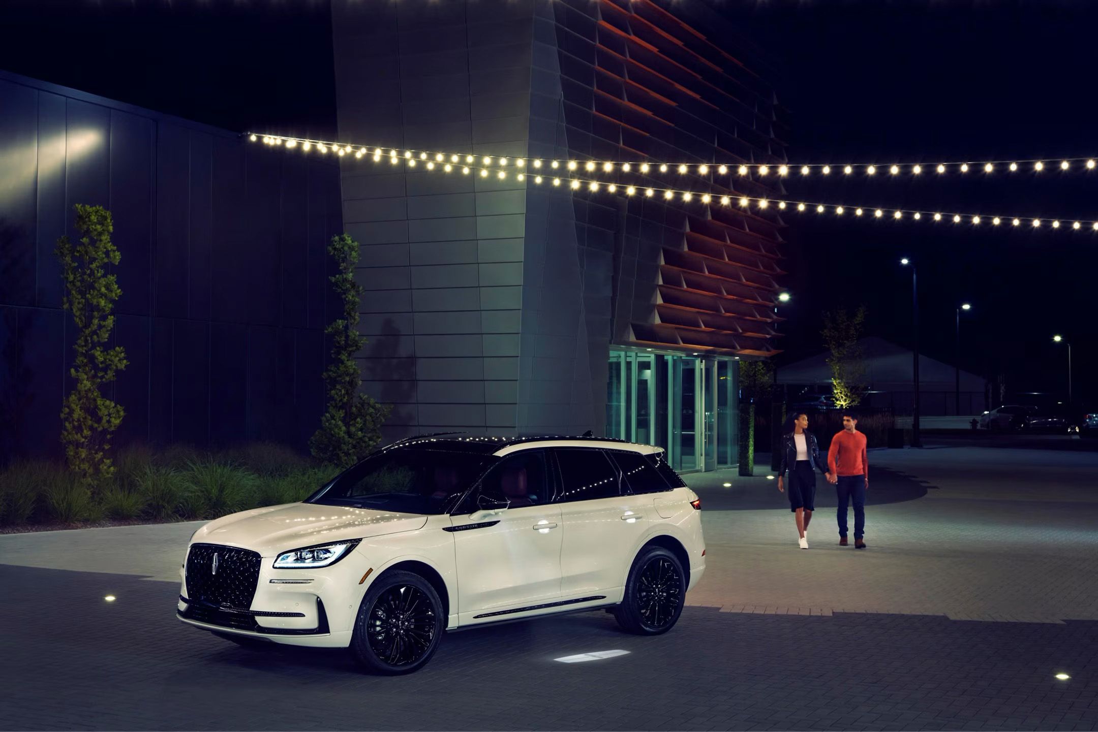 A white Lincoln Corsair SUV is parked at night in front of a modern building with string lights overhead. A couple is seen walking behind the vehicle, away from the building.