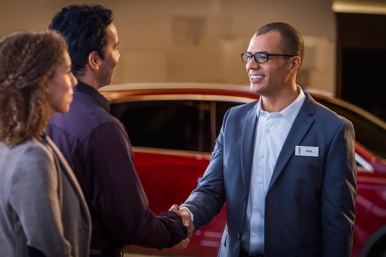 A man in glasses and a suit, wearing a "Phil" name tag, is shaking hands with another man. A woman with curly hair stands next to the man shaking hands, and a red car is visible in the background.
