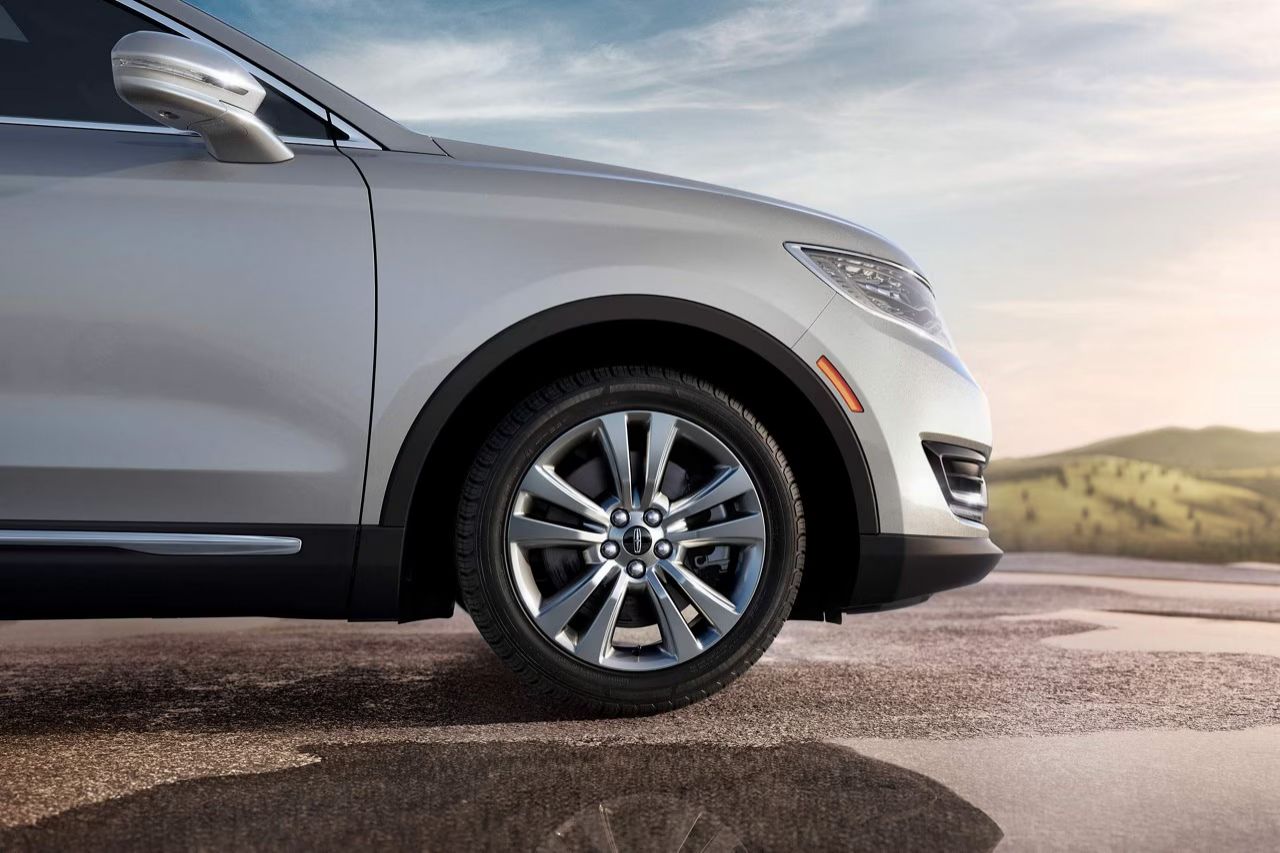  Close-up of the front tire and bumper of a silver Lincoln SUV parked on a wet surface, with rolling hills and a bright sky in the background.