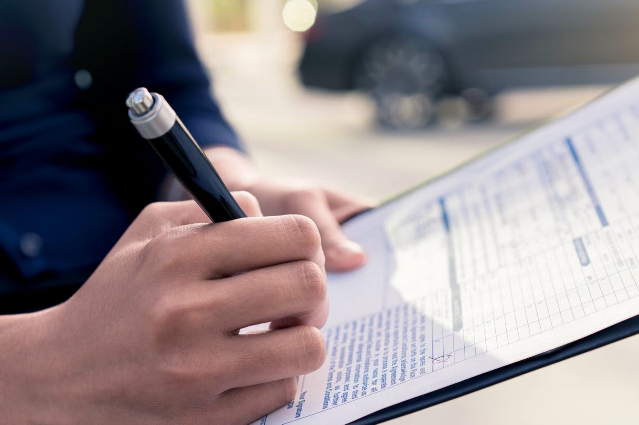  Close-up of a person's hands holding a pen and writing on a document or form, with a blurred car in the background.