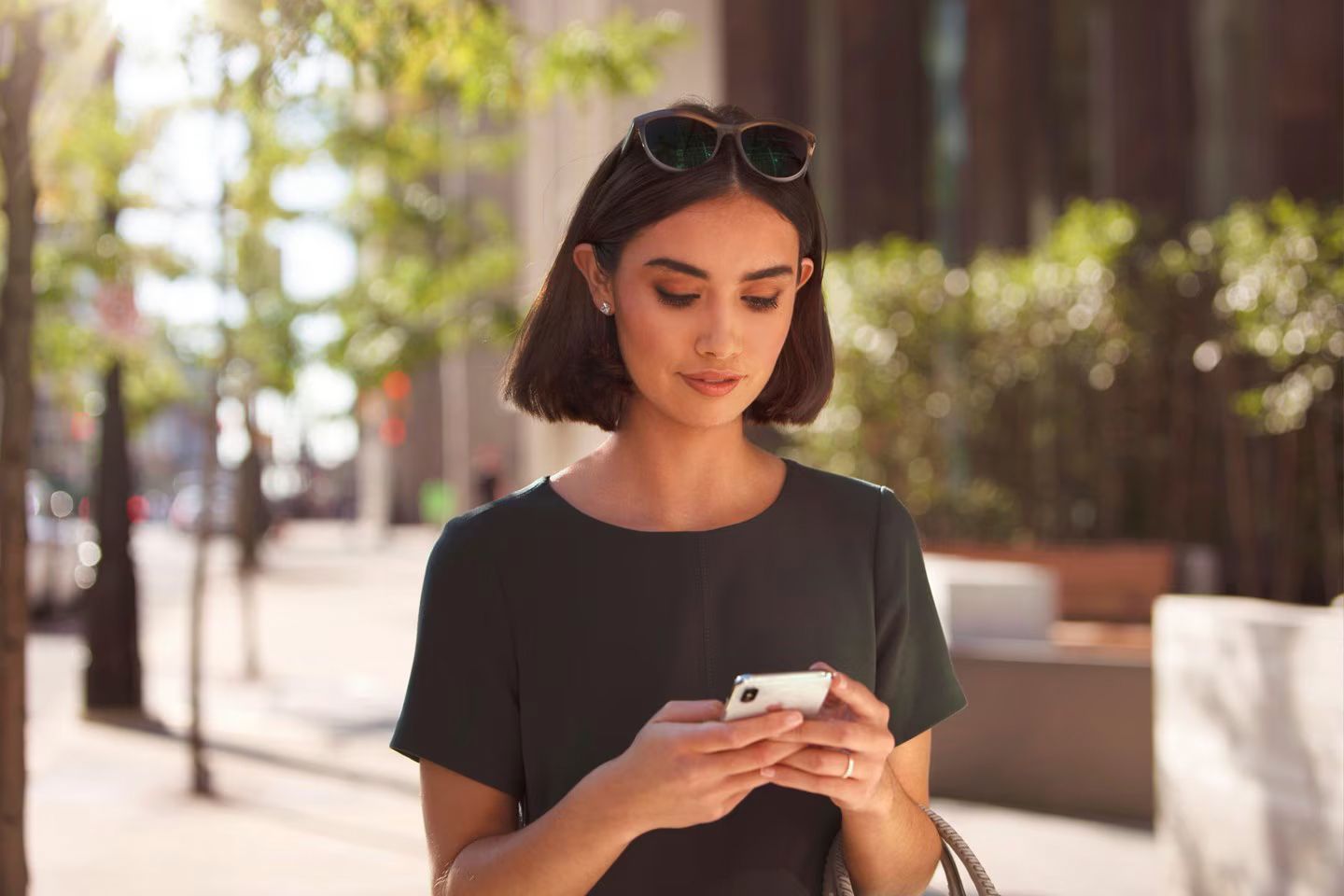A young woman with short, dark hair, wearing sunglasses on her head and a dark dress, is walking outdoors and looking at her cell phone.