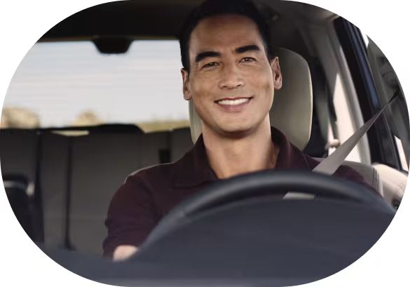 A man smiling directly at the camera while seated in the driver's seat of a car with his seatbelt on.