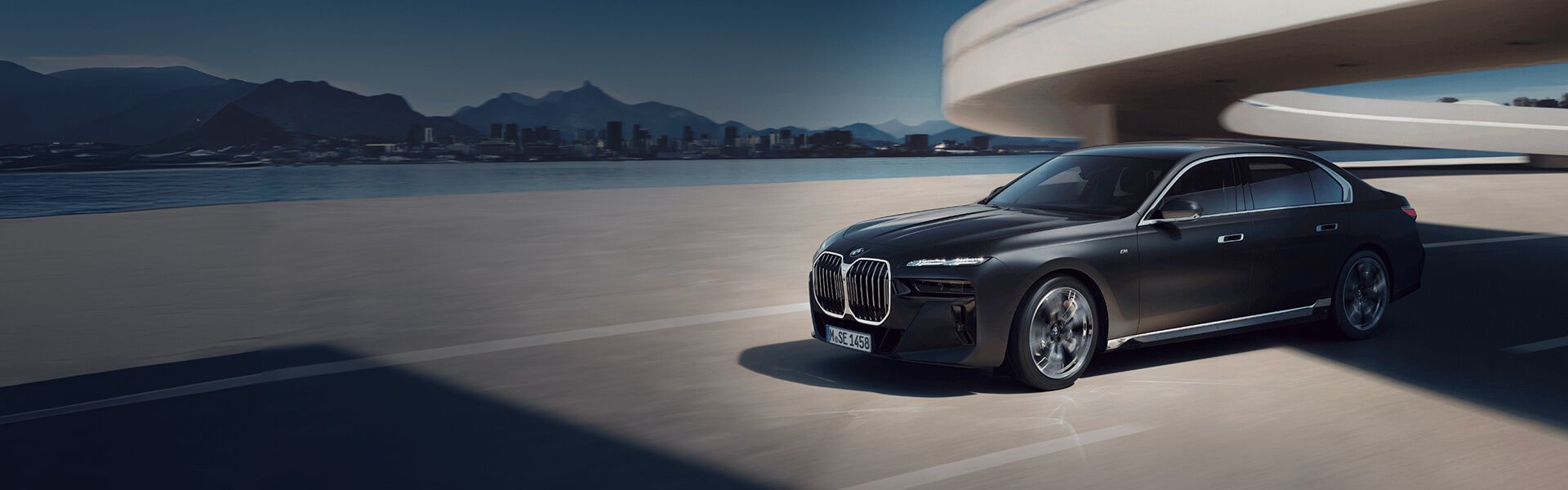 Large bannière d'une berline BMW Série 7 noire circulant sur une route côtière avec une silhouette urbaine et des montagnes en arrière-plan.