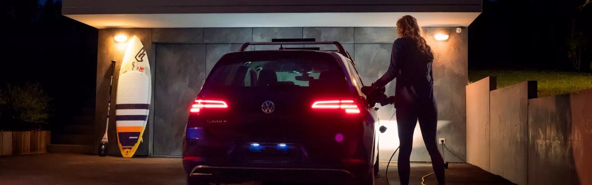 Une personne rechargeant une voiture électrique Volkswagen devant un garage la nuit, à côté d'une planche de surf.