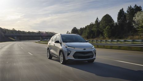 A white Chevrolet Bolt EUV driving along a highway lined with trees under a cloudy sky.