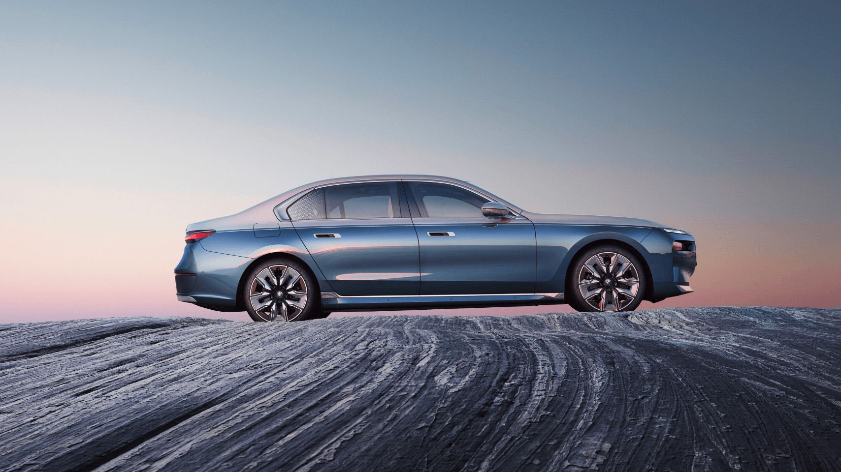 Side profile view of a light blue BMW 7 Series sedan parked on a textured rocky surface during sunset.