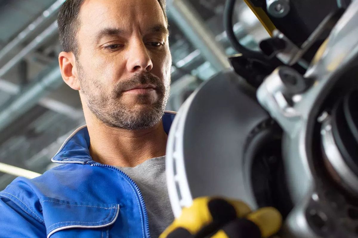 Gros plan d'un technicien automobile professionnel en uniforme bleu inspectant attentivement un composant de frein à disque dans un atelier.