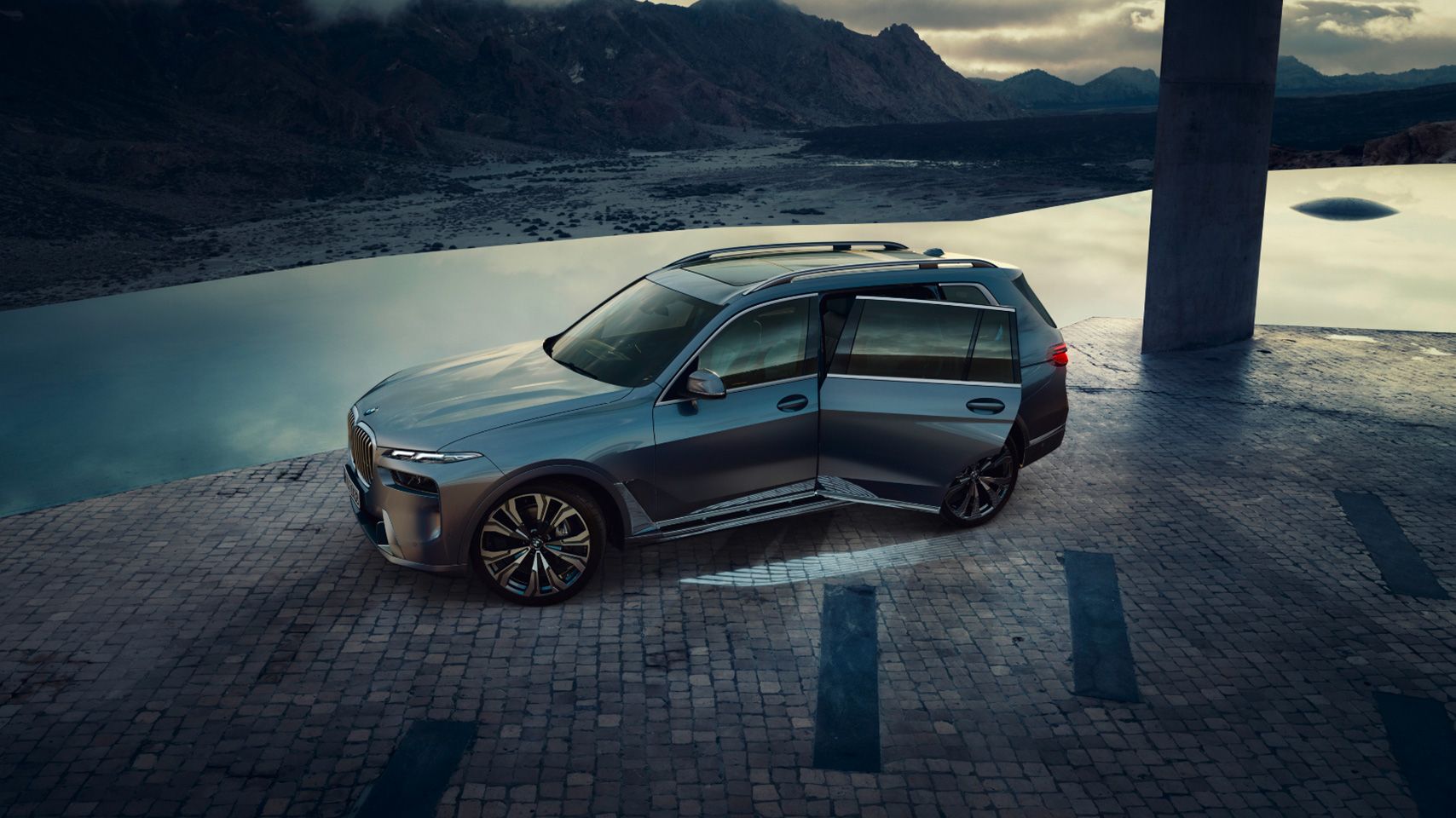 Exterior side view of a BMW X7 SUV with its doors open on a stone platform near water.