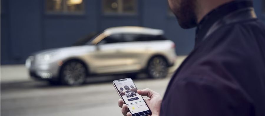 A detailed view from over a man's shoulder as he uses the Lincoln Way app on his smartphone, which displays a silver SUV. In the blurred background, a silver Lincoln luxury SUV is parked on a city street.