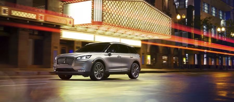Silver Lincoln luxury SUV parked on a city street at night under an illuminated theater marquee with red light trails streaking past