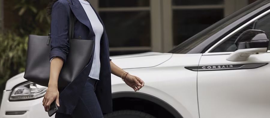 Woman carrying a tote bag and smartphone walking past a white Lincoln Corsair SUV