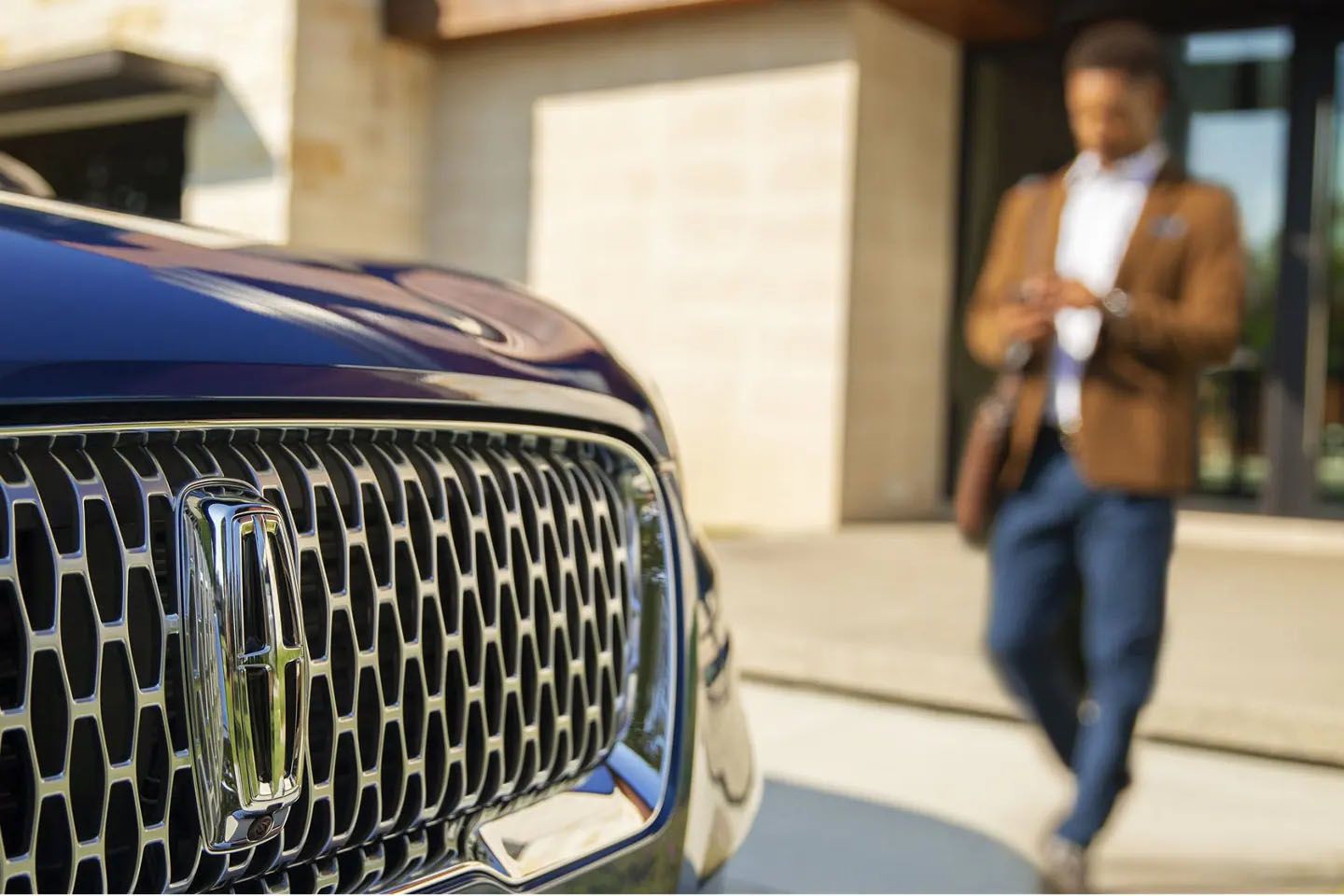 A detailed close-up of a dark blue Lincoln car's intricate chrome front grille with its distinctive logo. In the blurred background, a man in a brown blazer looks down at his smartphone.