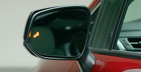Gros plan sur le rétroviseur extérieur d'un véhicule Toyota rouge, montrant l'icône lumineuse orange du moniteur d'angles morts activée.
