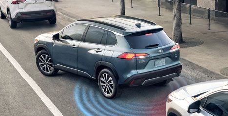 Vue en plongée d'un Toyota Corolla Cross bleu stationnant parallèlement, avec des ondes graphiques bleues et rouges illustrant les capteurs de recul et d'aide au stationnement.