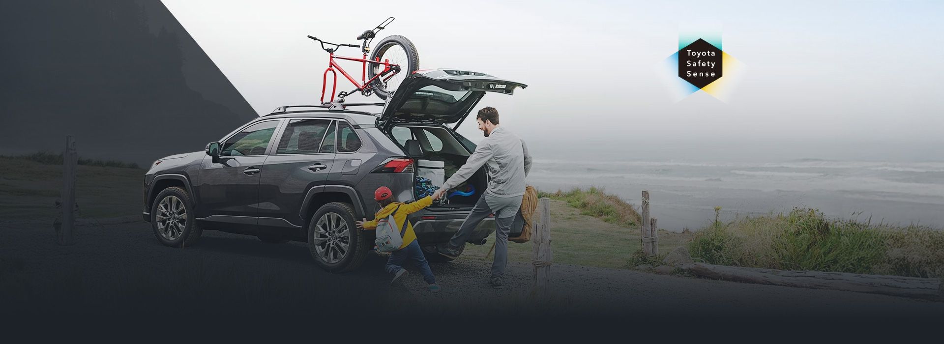 A family prepares for adventure next to their grey Toyota RAV4 equipped with a bike rack, featuring the Toyota Safety Sense logo against a misty coastal backdrop.