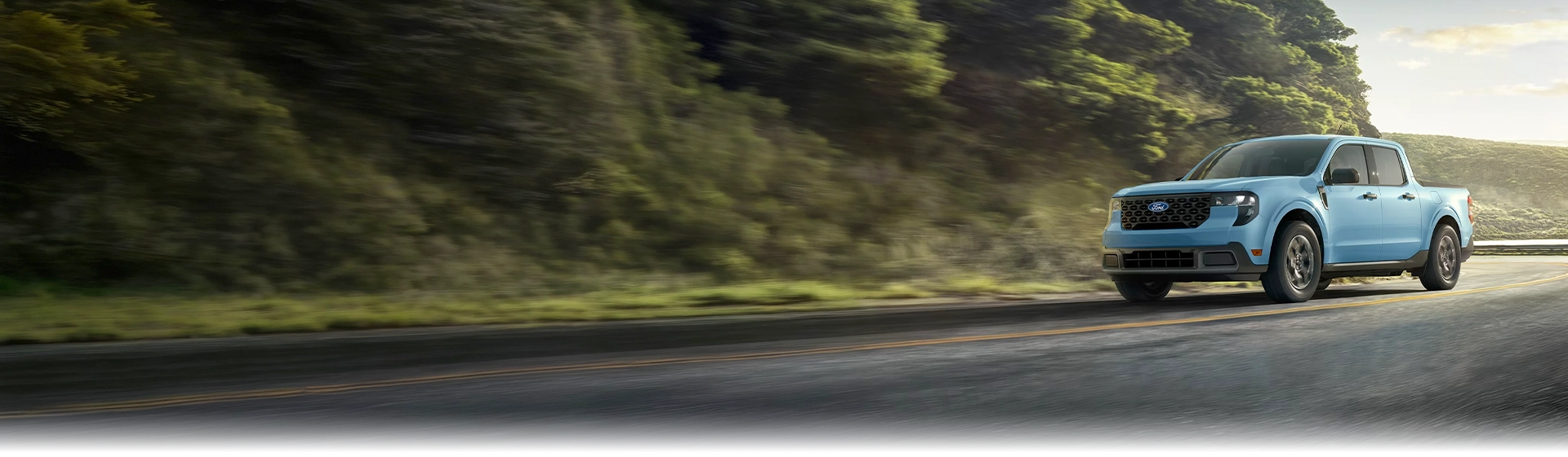 Light blue Ford Maverick pickup truck driving on a mountain road lined with trees.