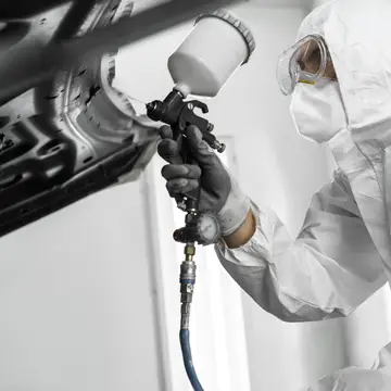 Technician in a protective suit spray painting a vehicle body part.