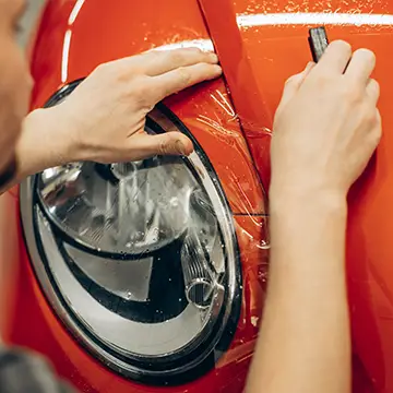 Applying a clear paint protection film onto a red car's headlight area.