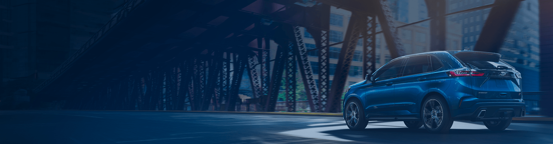 A blue Ford Edge ST SUV driving through a city street under an elevated bridge.