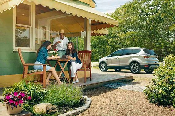Three friends relaxing on a porch looking at a tablet together, with a silver SUV parked in the background outside a house