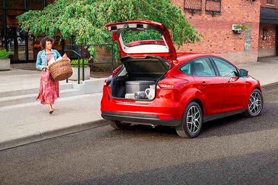 A woman carrying a basket walking toward a red hatchback car with its trunk open on a city street.