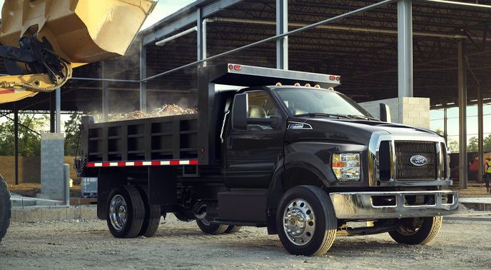 Black Ford commercial dump truck being loaded on a construction site, in front of an industrial structure.