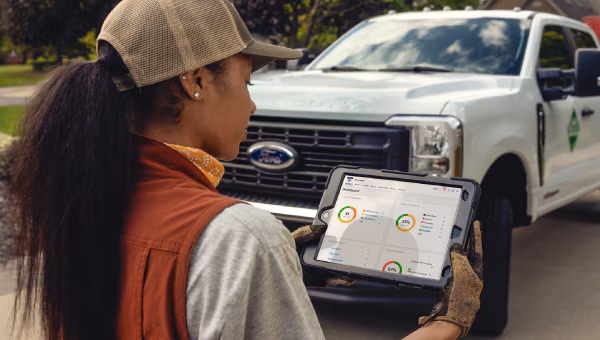 A female worker wearing a cap looks at a tablet displaying a Ford Pro fleet management dashboard in front of a white truck.